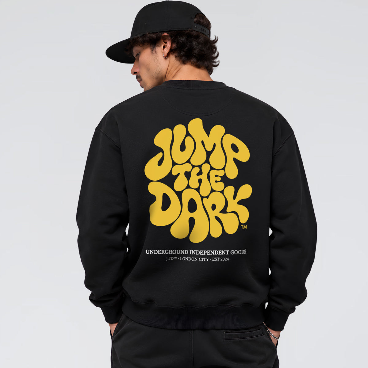 A man wearing a black crewneck sweatshirt from the back, featuring a large yellow retro-style graphic that reads 'JUMP THE DARK' with 'UNDERGROUND INDEPENDENT GOODS' printed below it. 